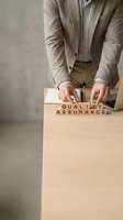 Caucasian male professional in beige blazer and white shirt arranging wooden letter blocks spelling quality assurance on desk with laptop, representing business standards and compliance.