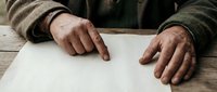 Weathered elderly male hands pointing at blank paper on rustic wooden table, symbolizing writing, signing, or reading concepts.