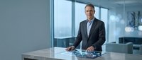 Professional middle-aged Caucasian businessman in dark suit standing confidently at modern glass desk in contemporary office with city view windows.
