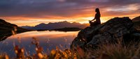 Woman sitting on rocky outcrop overlooking serene mountain lake at sunset, golden hour landscape with dramatic sky, peaceful nature scene for travel and wellness content.