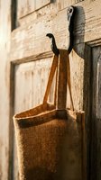 Rustic burlap tote bag hanging on vintage wooden door hook with warm sunlight creating natural shadows on textured brick wall background.