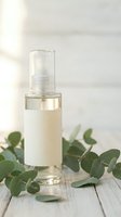 Clear glass spray bottle with natural serum on white wooden surface surrounded by fresh eucalyptus leaves for organic skincare and wellness products.