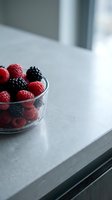 Fresh red and black raspberries in glass bowl on modern kitchen countertop with natural window light, suitable for food photography, healthy eating, and culinary content.