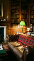 Cozy library study room with leather Chesterfield sofa, red tartan throw, green desk lamp, wooden bookshelves, fireplace, and warm ambient lighting for interior design and home decor projects.