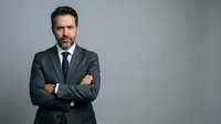 Middle-aged man with dark hair and beard wearing charcoal business suit and navy tie, arms crossed, confident professional portrait against neutral gray background.