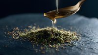 Golden honey dripping from spoon onto dried herbs and spices scattered on dark stone surface, creating artistic food composition in moody lighting.
