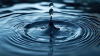 Macro photography of water drop impact creating concentric ripples and crown splash formation in dark blue tones, captured with high-speed technique.