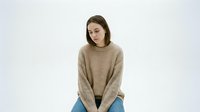 Young woman with long brown hair and fair complexion wearing a beige ribbed sweater and blue jeans, sitting against white background for portrait or fashion use.
