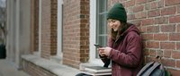 Young woman in burgundy hoodie and green beanie using smartphone while sitting outdoors against brick building wall, casual urban lifestyle and technology.