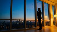 Professional businessman silhouette standing by floor-to-ceiling windows overlooking illuminated city skyline at dusk in modern corporate office interior.