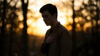 Young man with dark hair and olive complexion standing outdoors at golden hour sunset, hand on chest in contemplative pose among bare trees, warm backlit lighting.