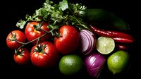 Fresh vegetables and fruits on black background with water drops: tomatoes on vine, red chili peppers, limes, red onion, zucchini and cilantro arranged for food photography.