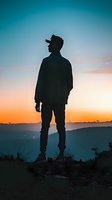 Silhouetted male hiker standing on mountain ridge at sunset, wearing cap and casual jacket, gazing at colorful sky. Outdoor adventure and travel concept.