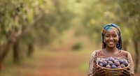 Young African woman holding basket of fresh plums in orchard, smiling at camera with fruit trees in background.