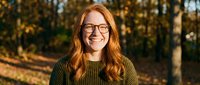 Happy young Caucasian woman with red hair and glasses smiling outdoors in autumn park with golden sunlight and blurred foliage background for lifestyle content.