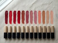 Lipstick color palette swatches arranged in gradient from deep red to nude shades with gold tubes on neutral fabric background for beauty testing.