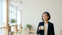 Young Asian woman in professional black blazer holding white coffee cup at desk with laptop and potted plants, suitable for business, corporate, and lifestyle content.