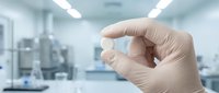 Gloved hand holding small pharmaceutical capsule in sterile laboratory environment with medical equipment and bright lighting.