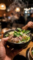 Steaming bowl of Asian noodle soup with beef slices, fresh herbs, and vegetables held in hand at restaurant with warm ambient lighting.