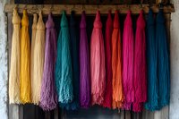 Vibrant multicolored yarn skeins hanging on wooden rack against rustic wall, featuring yellow, purple, turquoise, pink and blue tassels in traditional textile display.