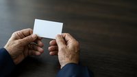 Senior male hands holding blank white business card on dark wooden surface for corporate branding and professional networking purposes.