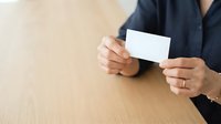 Professional woman in dark blazer holding blank white business card at wooden table, suitable for corporate branding, business identity, and professional networking materials.