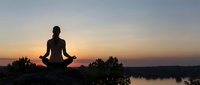Woman silhouette meditating in lotus pose at sunset by lake. Peaceful yoga practice during golden hour with serene water reflection and tranquil sky.