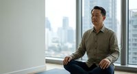 Middle-aged Asian businessman meditating in modern office space by window with city view, practicing mindfulness and stress relief during workday.