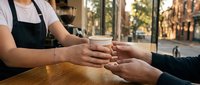 Barista or cafe worker with medium skin tone wearing black apron serving coffee to customer at wooden counter, hands exchanging cup in bright cafe setting with window view.