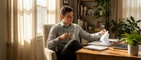 Young professional man reviewing documents while drinking coffee in bright home office with plants and natural lighting for remote work productivity.