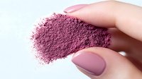 Crushed pink mineral makeup powder pigment held between fingers with matching manicure on white background, showing cosmetic texture and application.