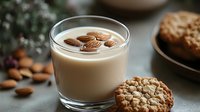 Creamy almond milk with floating nuts in glass cup served with homemade oatmeal cookies on gray background, perfect for breakfast or snack.