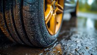 Close-up view of wet car tire with golden alloy wheel on rainy asphalt road, showing tread pattern and water droplets in automotive setting.