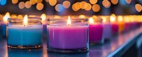 Colorful aromatic candles in glass jars with soft bokeh lights background create cozy atmosphere. Blue and purple wax glows with warm flames in panoramic view.