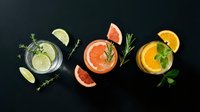 Three colorful cocktails with fresh citrus garnishes and herbs arranged on dark background for bar menu design and beverage photography.