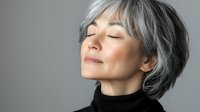 Asian mature woman with silver gray hair and black turtleneck sweater expressing serenity and mindfulness with closed eyes against neutral background.