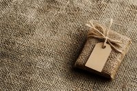 Small gift wrapped in burlap with twine bow and blank kraft paper tag on natural jute background, rustic eco-friendly packaging.