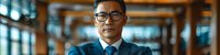 Confident Asian businessman in glasses and navy suit standing with arms crossed in modern office corridor, professional corporate portrait.