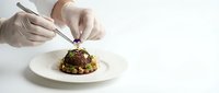 Professional chef hands in white gloves carefully plating gourmet dish with tweezers on white plate against clean background for culinary presentation.