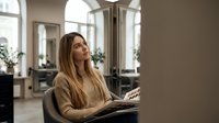 Young blonde businesswoman reading magazine in modern office interior during break time relaxing in comfortable chair near large windows.