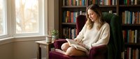 Young woman reading book in cozy library armchair surrounded by bookshelves with warm natural light streaming through window creating peaceful study atmosphere.