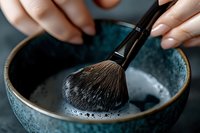 Makeup brush being cleaned in ceramic bowl with soapy water and foam. Close-up view showing soft bristles and female hands with manicure washing cosmetic tool.