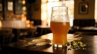 Craft beer glass filled with golden ale on wooden bar counter with hops and wheat, warm pub atmosphere with bokeh lights in background for brewery marketing.