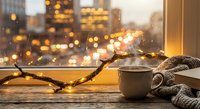 Cozy winter evening scene with warm coffee mug, knitted scarf and glowing string lights on wooden windowsill overlooking blurred city lights at dusk.