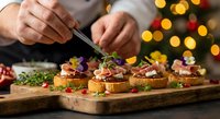 Chef preparing gourmet holiday appetizers with tweezers on wooden board against festive Christmas lights background for catering events.