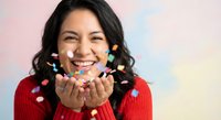 Young Hispanic woman in red sweater blowing colorful confetti with joyful expression against soft pastel background for celebration and party concepts.