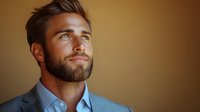 Confident young Caucasian businessman with beard wearing grey suit jacket and blue shirt looking away thoughtfully against warm brown background in professional portrait.
