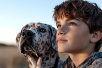 Young boy with curly hair and Dalmatian dog looking up at sunset, capturing bond between child and pet in warm golden light.