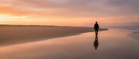 Solitary figure standing on wet sand beach at sunset, casting reflection in shallow water during golden hour, serene coastal landscape.