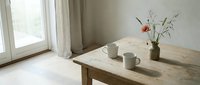 Minimalist wooden dining table with ceramic mugs and flower vase in bright natural light interior. Scandinavian home decor style for kitchen design projects.